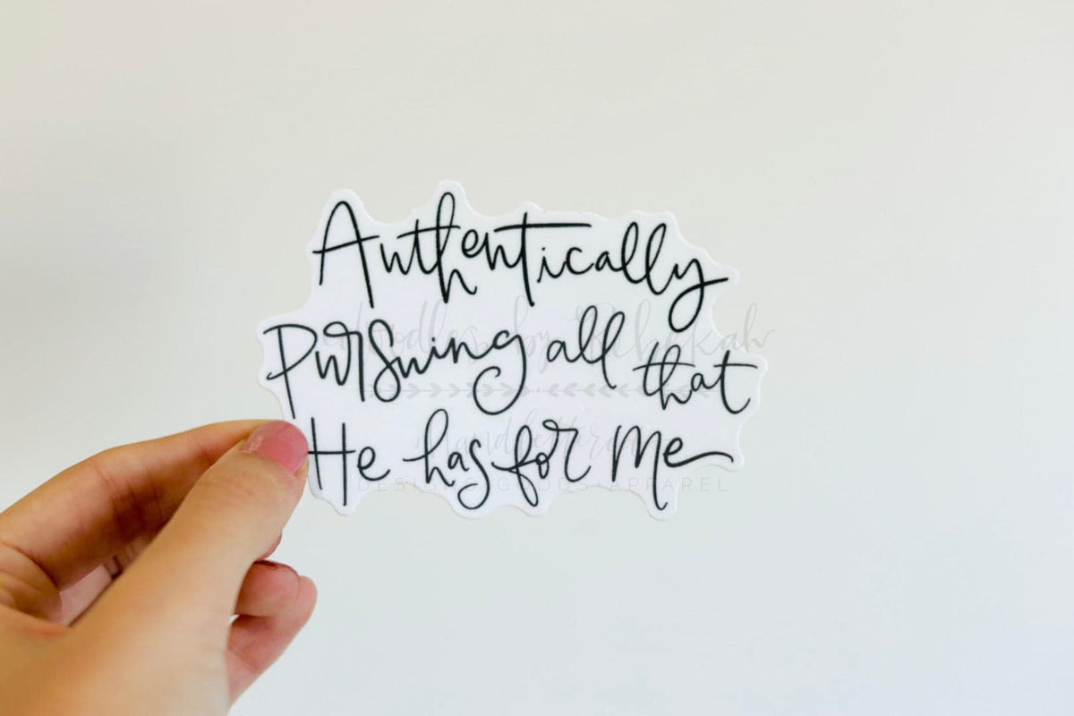 A hand holding a decorative sticker with the text 'Authentically Pursuing All That He Has For Me' written in cursive script.