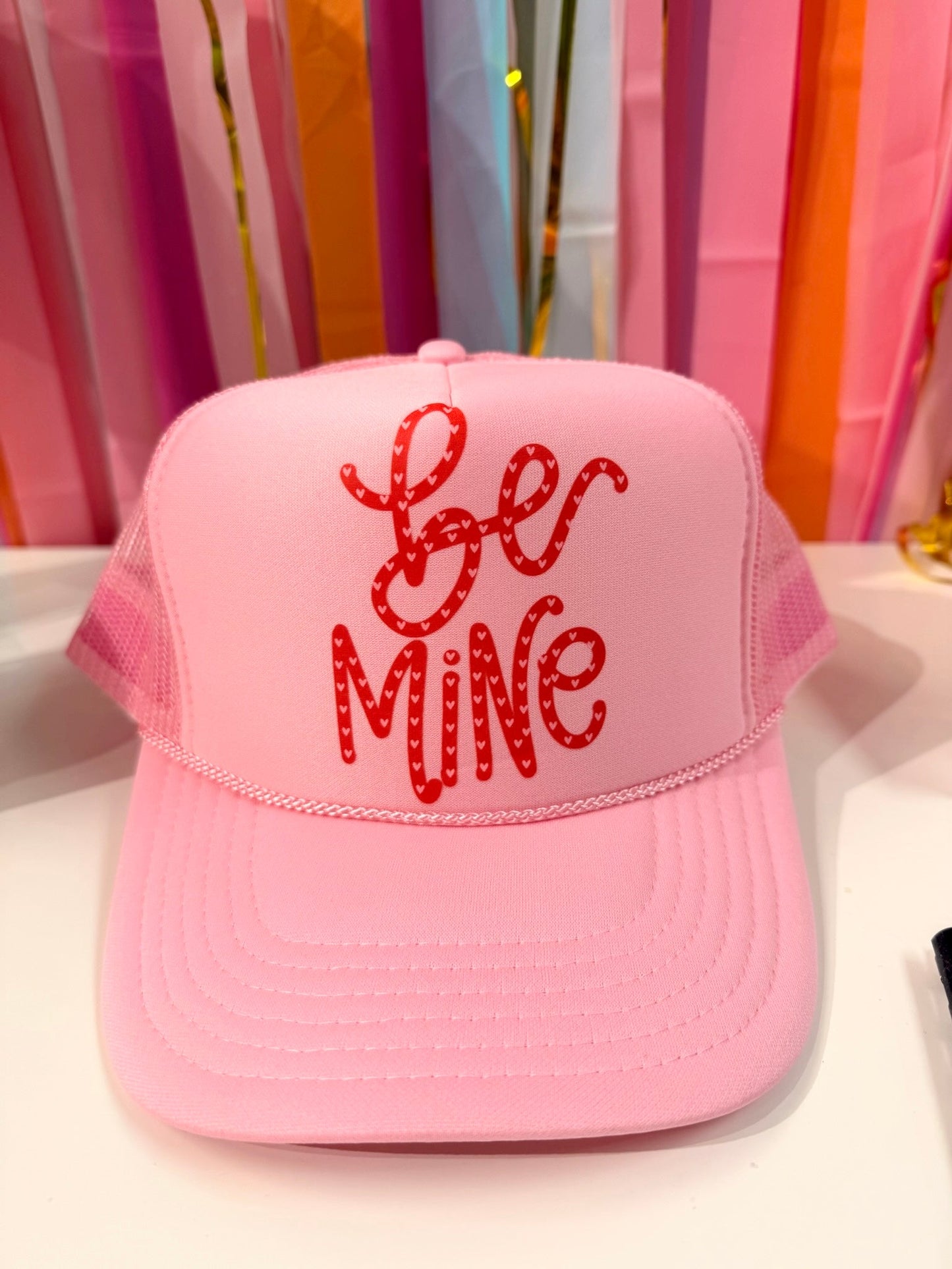 Doodles by Rebekah "Be Mine" Valentine's Day trucker hat featuring whimsical hand-drawn typography with heart patterns on a colorful party streamer background.