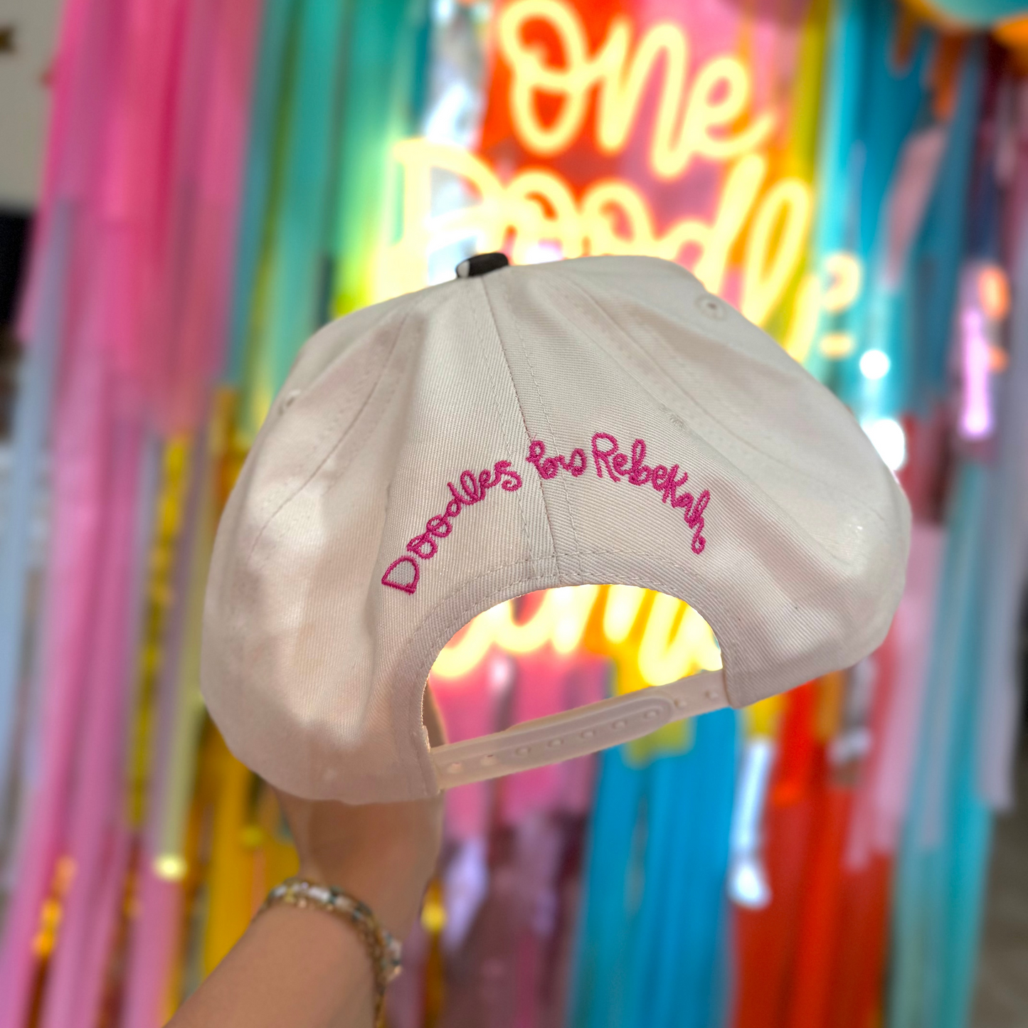 White cap with pink text held in front of colorful balloons and a neon sign.