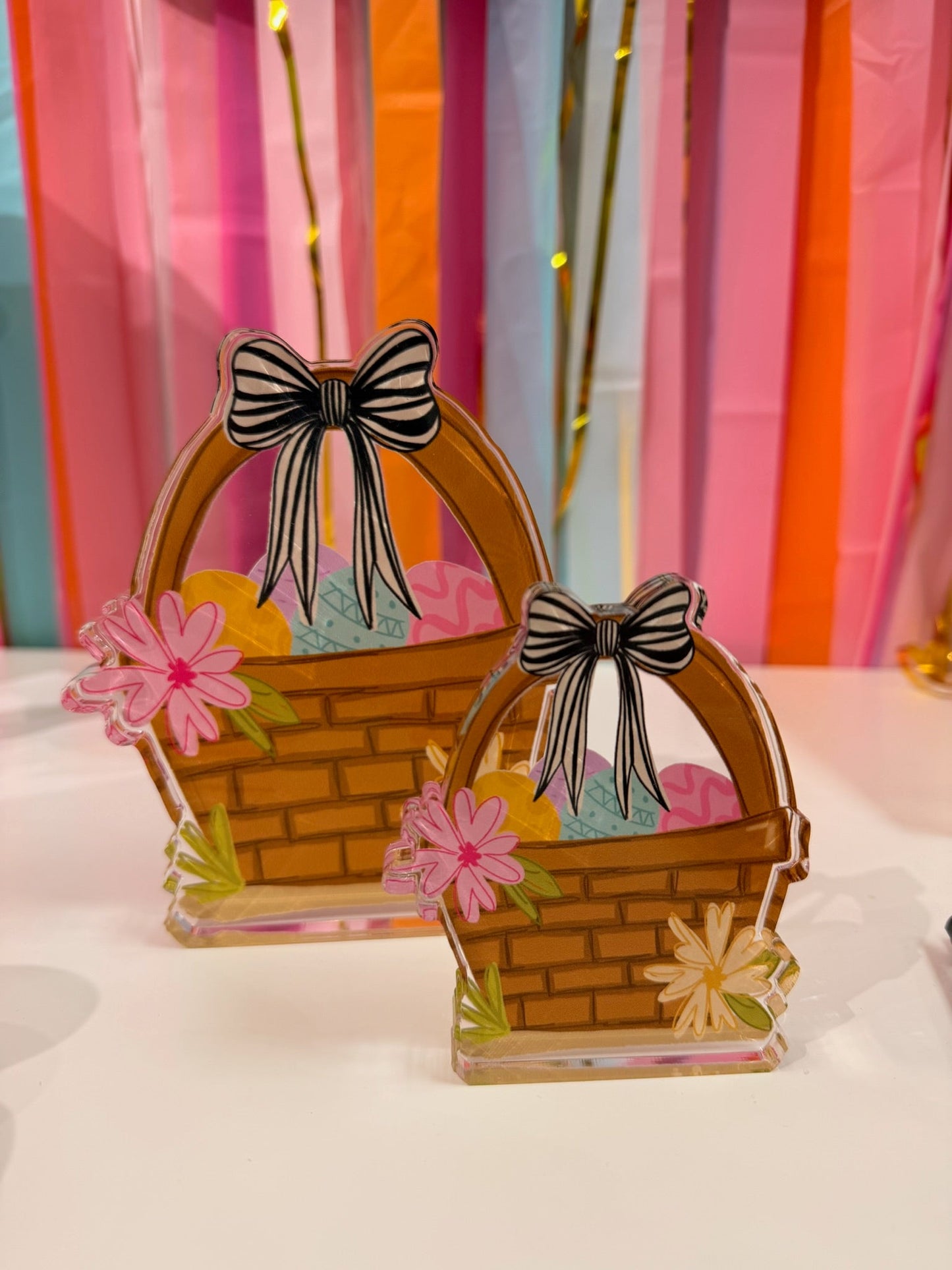 Doodles by Rebekah Easter basket acrylic home decor featuring whimsical hand-painted eggs, a striped bow, and floral details. These colorful spring tabletop accents are shown against a bright party streamer background.