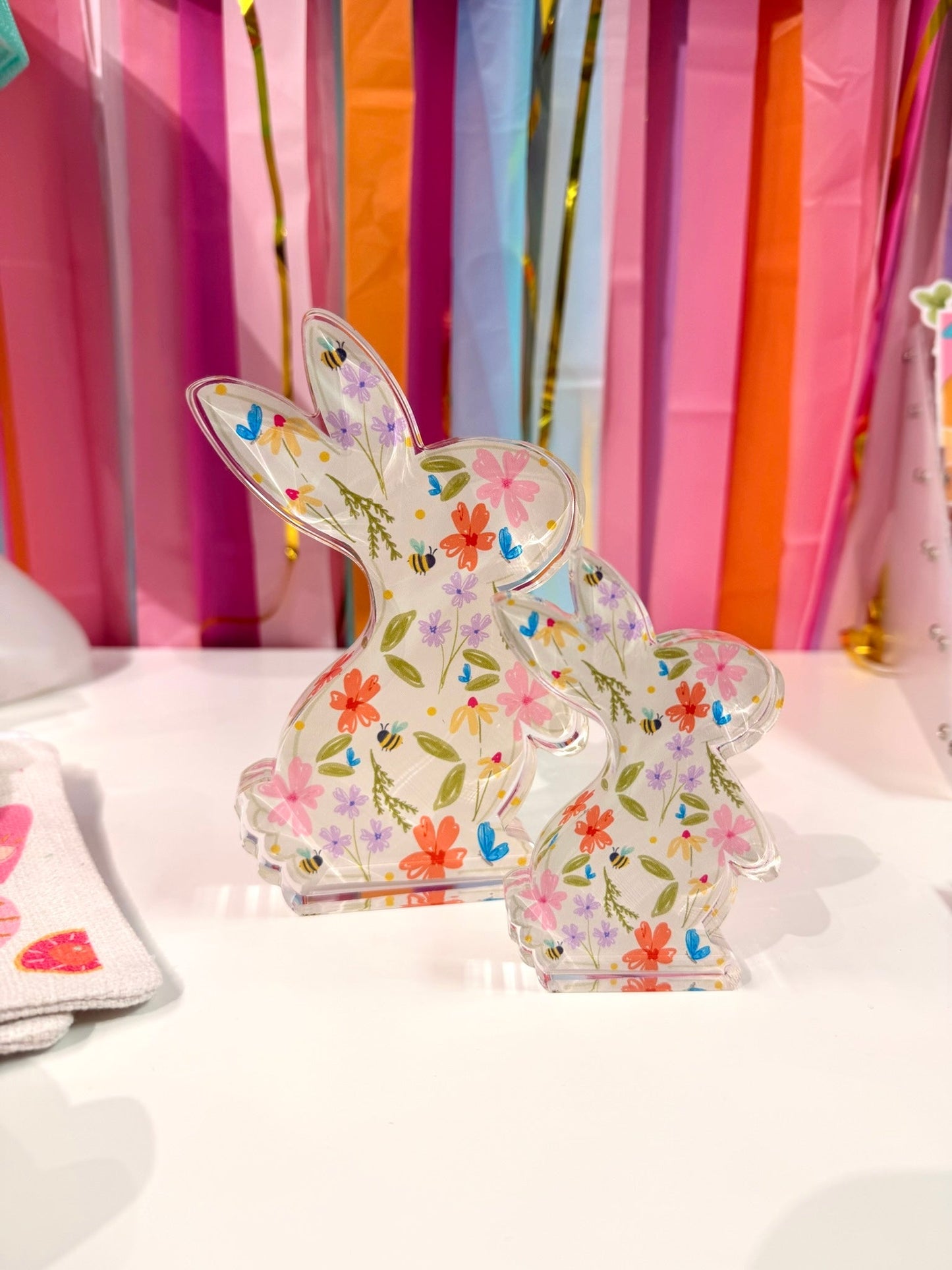 Doodles by Rebekah floral bunny acrylic home decor featuring whimsical hand-painted spring wildflowers and honeybee patterns. These colorful Easter tabletop decorations are shown against a bright party streamer background.