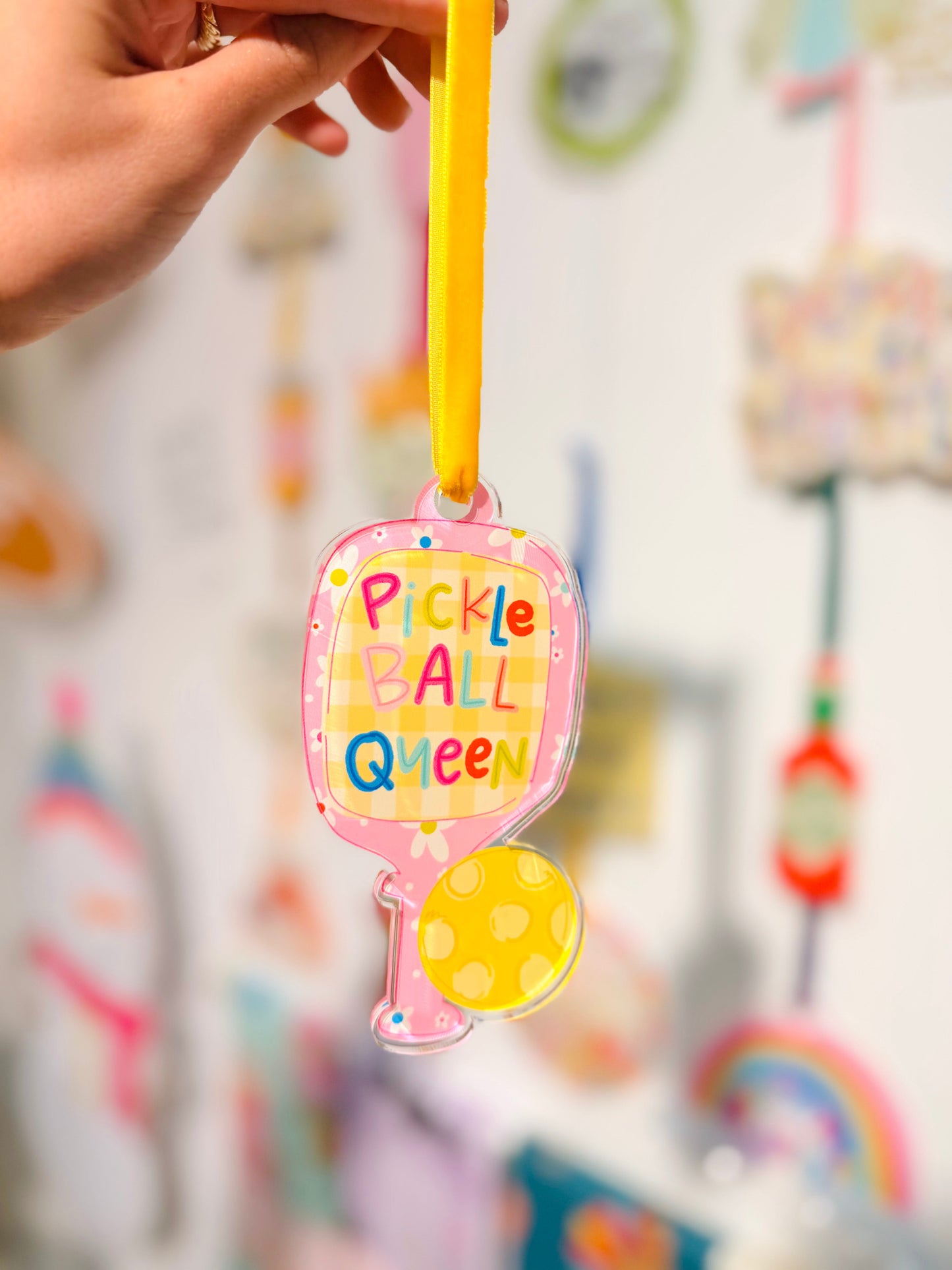 Colorful 'Pickle Ball Queen' ornament held by a hand with a blurred background of colorful items.