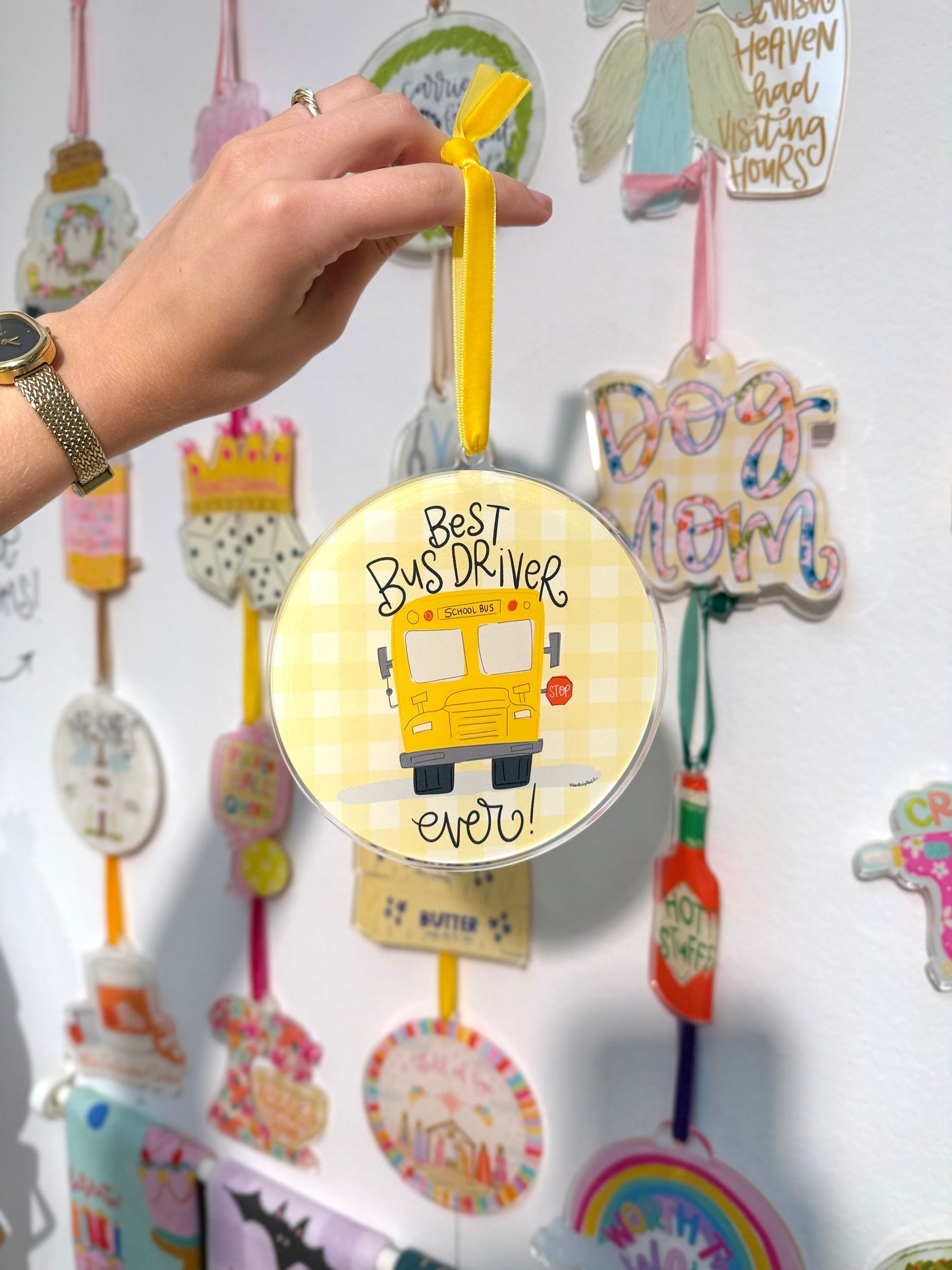 Hand holding a decorative ornament with 'Best Bus Driver ever!' text against a wall of other ornaments.