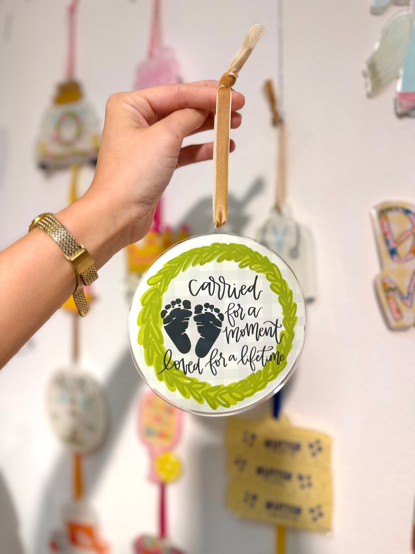 Hand holding a circular ornament with text and footprints, against a blurred background of similar ornaments.