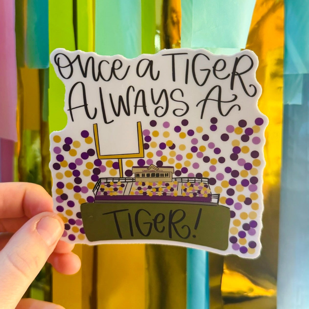 Doodles by Rebekah "Once a Tiger Always a Tiger" school spirit sticker. This hand-painted vinyl decal features a football stadium and purple and gold confetti, making it a perfect gift for students, alumni, and fans of tiger-mascot schools to display on water bottles and laptops.