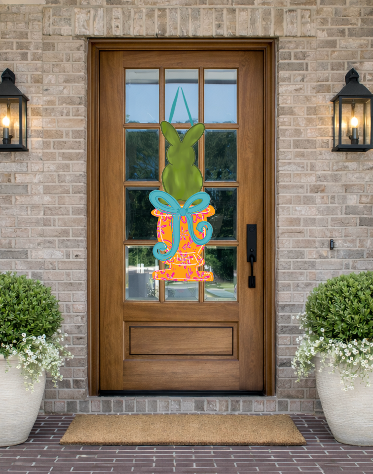 Doodles by Rebekah spring door decor featuring a whimsical green bunny in an orange floral urn with a blue bow, displayed on a rustic front porch.