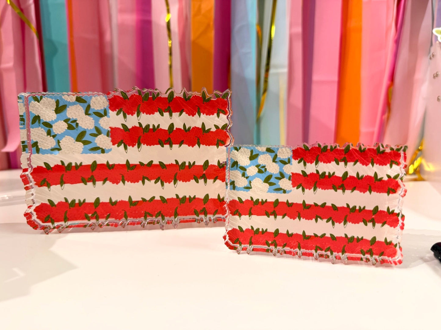 Doodles by Rebekah patriotic floral American flag acrylic home decor featuring whimsical hand-painted red flower stripes and white floral stars. These unique Fourth of July accents are shown against a bright party streamer background.