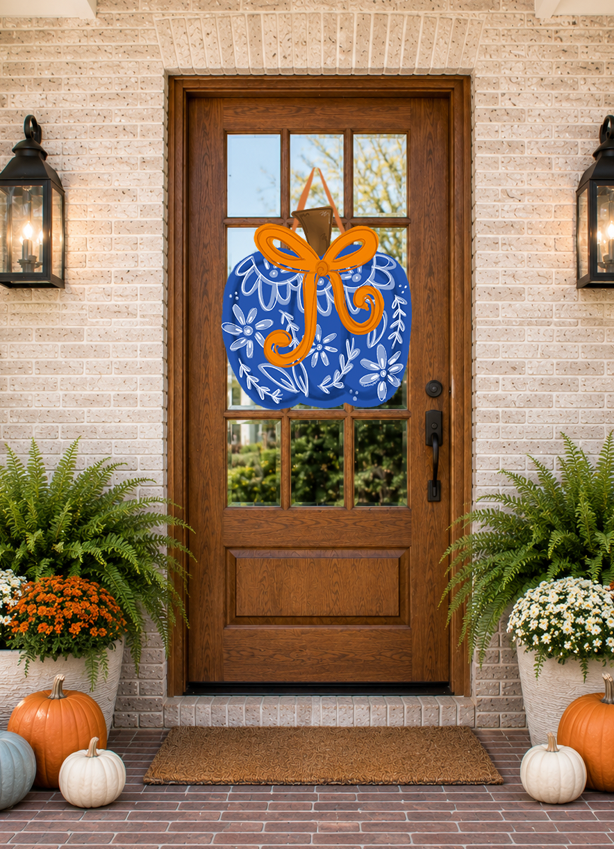 Decorative blue wreath with an orange bow on a wooden door with glass panels, flanked by potted plants and pumpkins.