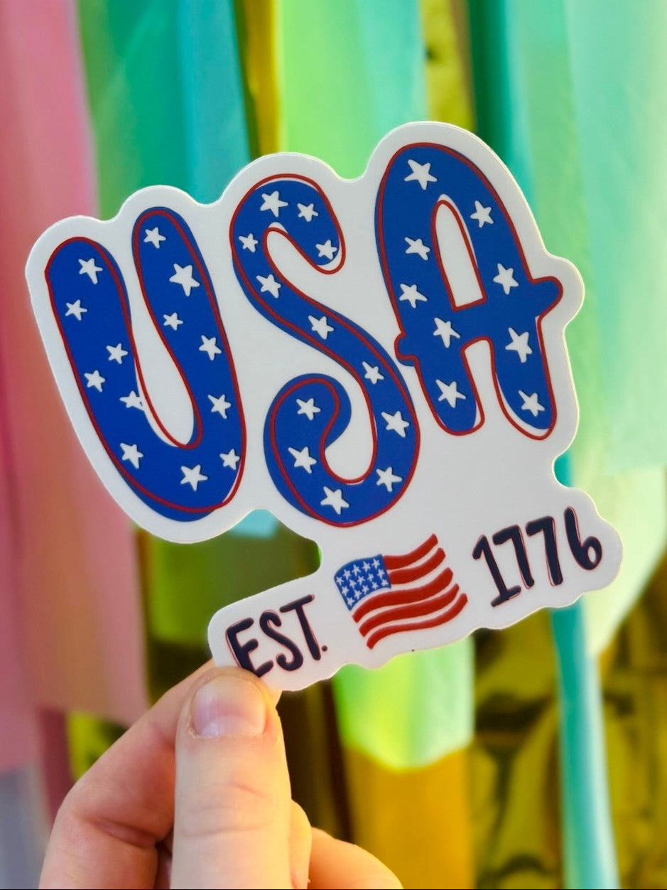 Person holding a USA sticker with "EST 1776" and a flag