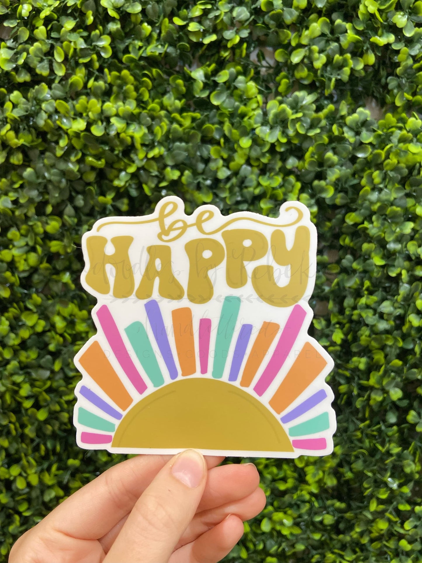 A sticker with a multicolored design featuring the text 'Be Happy' held in front of a green leafy background.