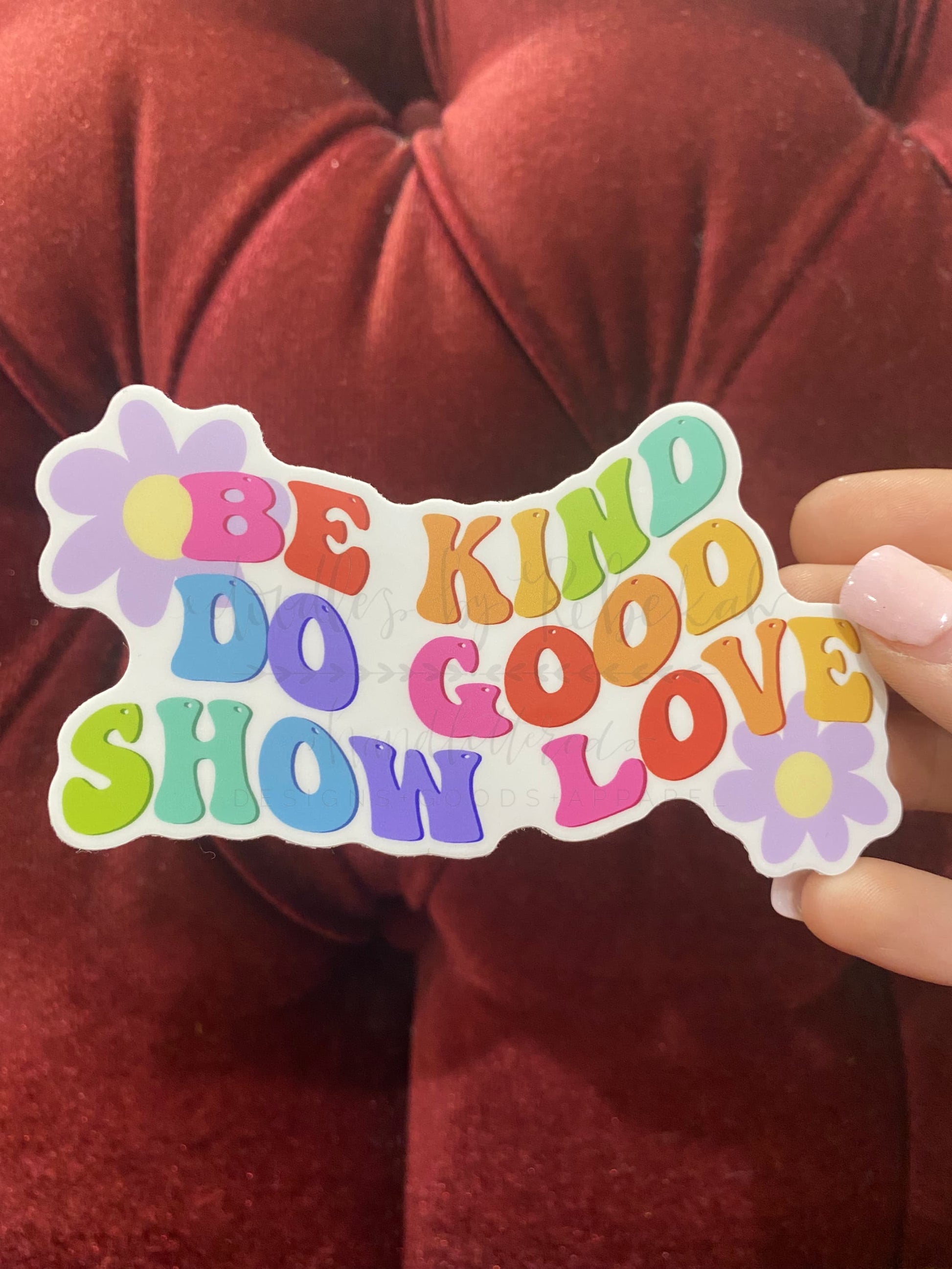A colorful sticker with the text 'BE KIND DO GOOD SHOW LOVE' written in a playful, multicolored font, held against a red background.