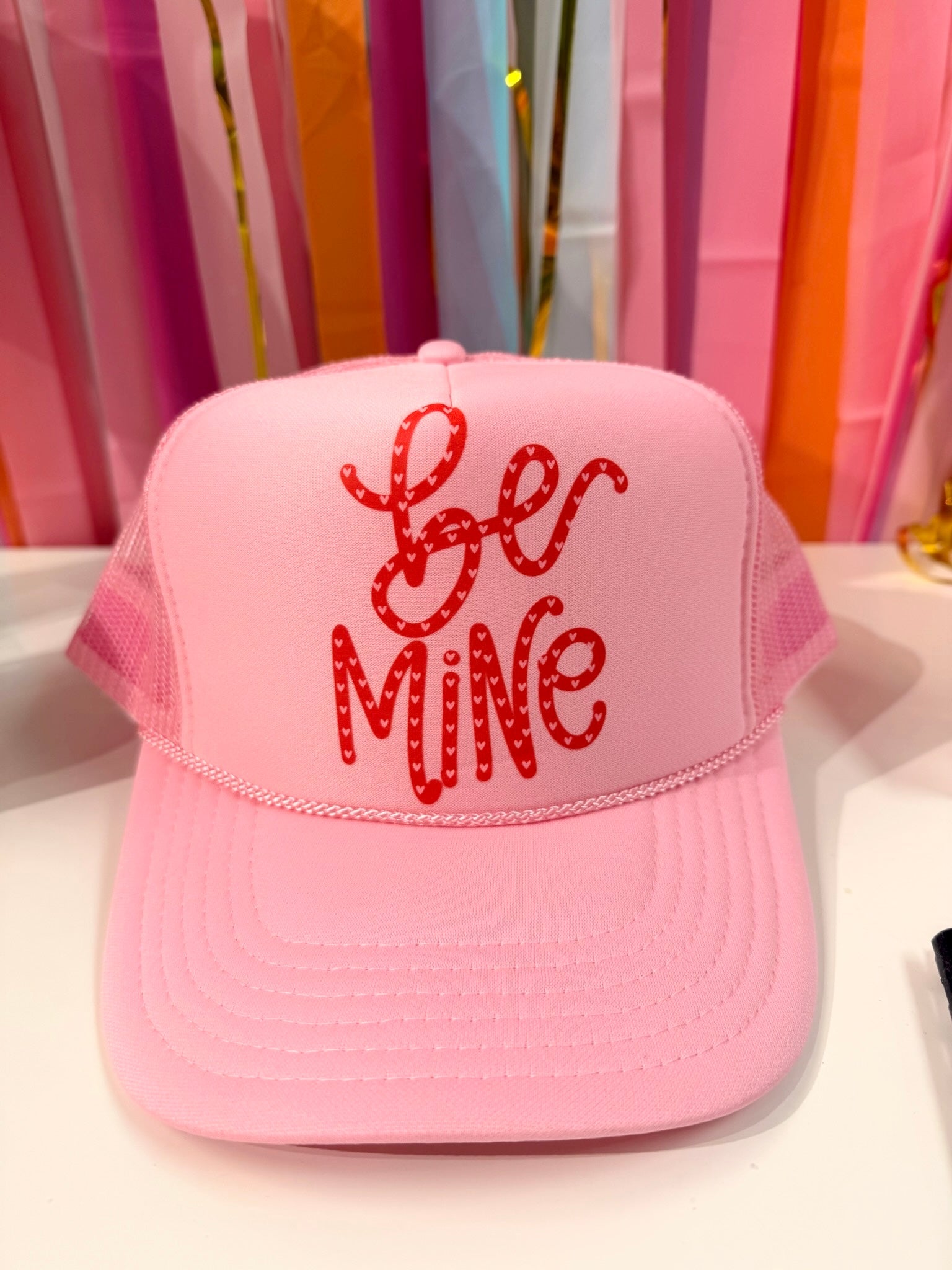 Doodles by Rebekah "Be Mine" Valentine's Day trucker hat featuring whimsical hand-drawn typography with heart patterns on a colorful party streamer background.