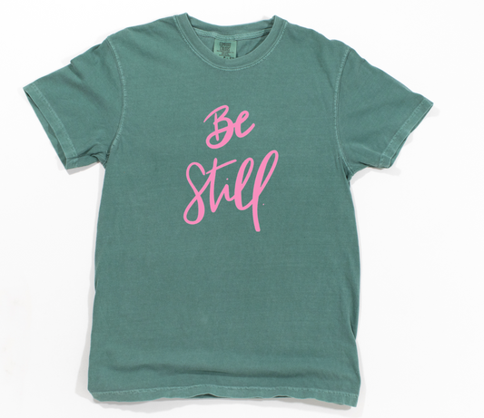 Green t-shirt with 'Be Still' in pink text on a white background