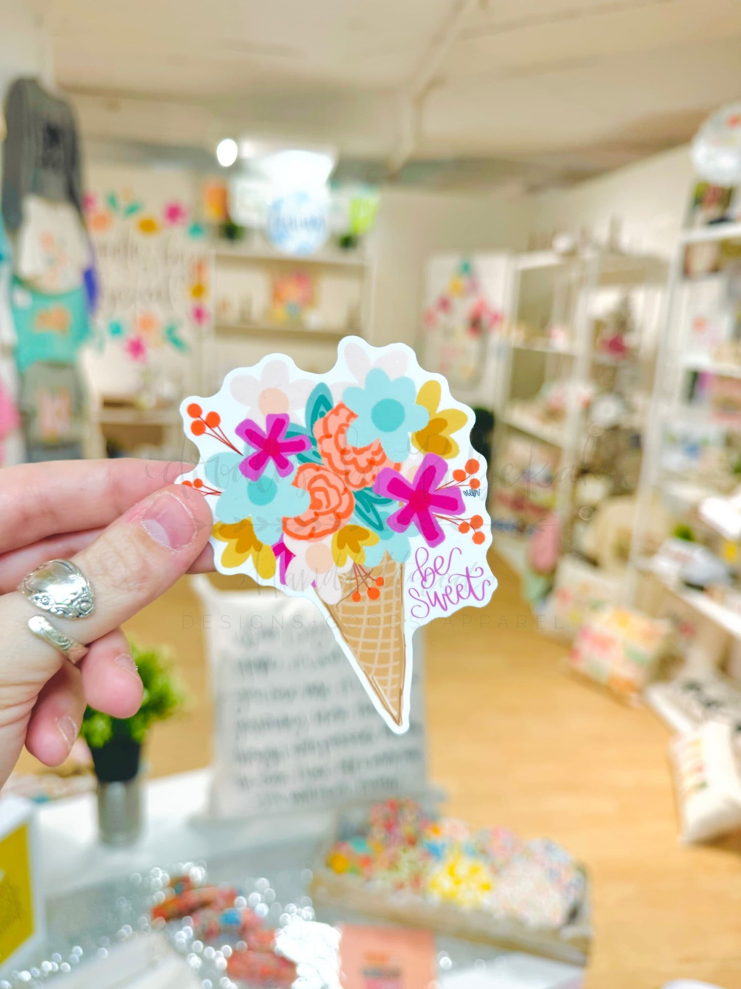 A sticker with a colorful floral design on a cone shape, held up against a blurred shop display background.