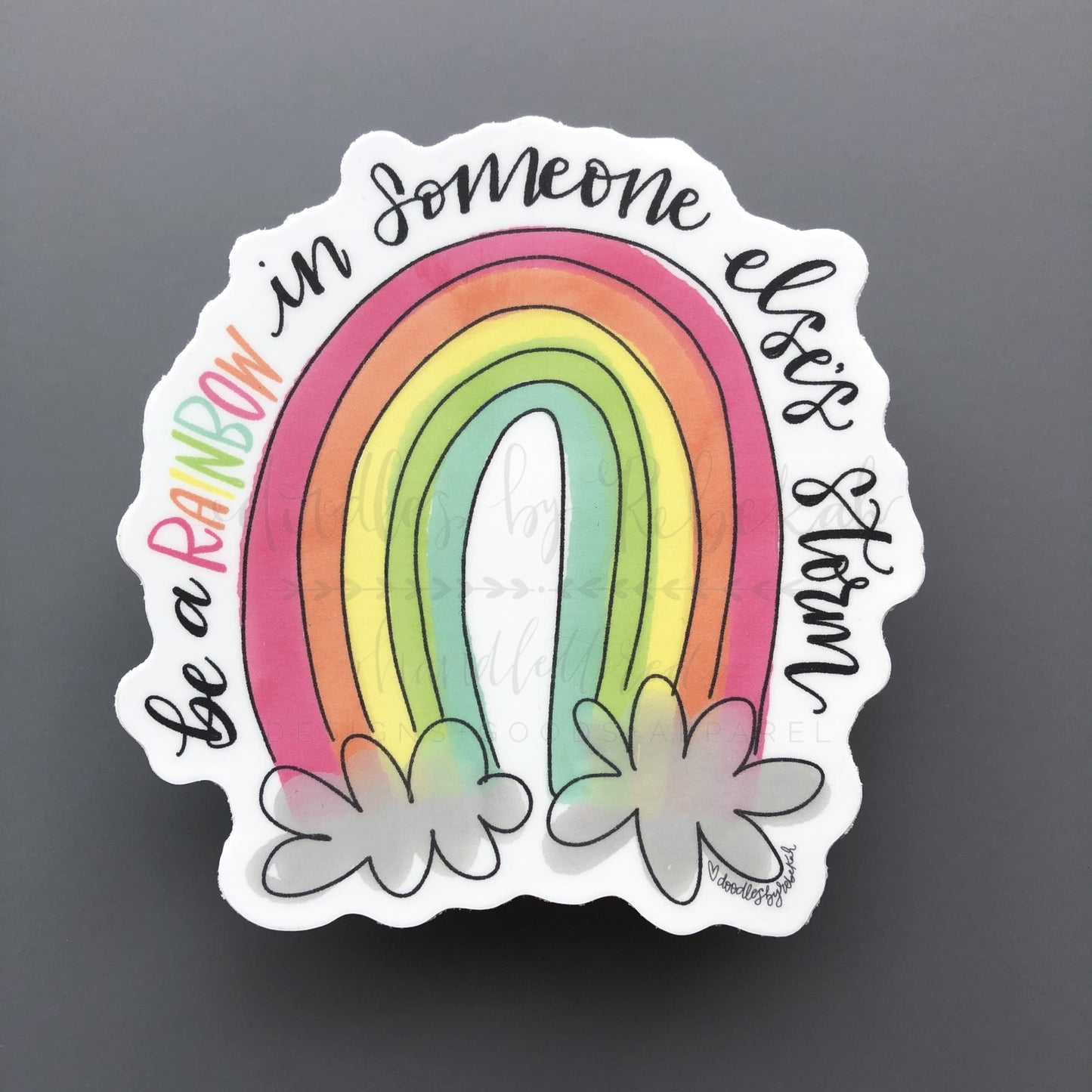 A sticker with a colorful rainbow design and text 'be a rainbow in someone else's storm' surrounded by flowers at the bottom.