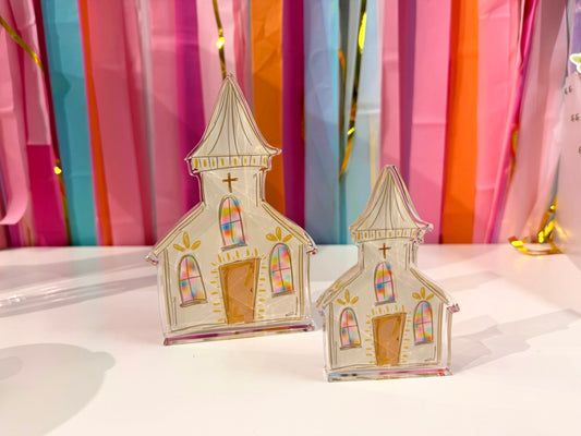 Doodles by Rebekah white church acrylic home decor featuring whimsical hand-painted stained glass windows and gold accents. These colorful religious or spring tabletop decorations are shown against a bright party streamer background.
