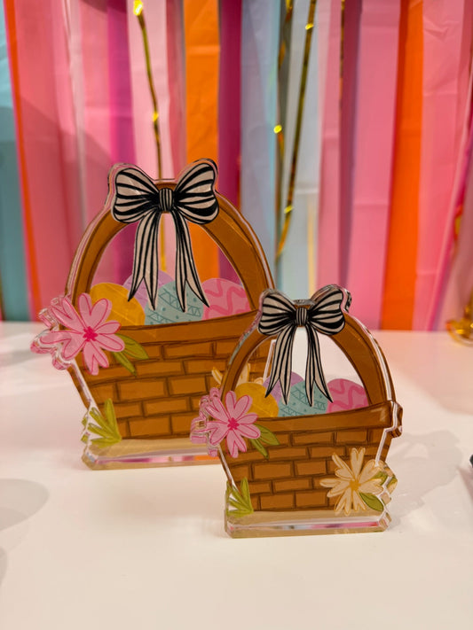 Doodles by Rebekah Easter basket acrylic home decor featuring whimsical hand-painted eggs, a striped bow, and floral details. These colorful spring tabletop accents are shown against a bright party streamer background.