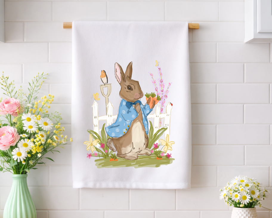 Whimsical Peter Rabbit inspired decorative kitchen tea towel featuring a bunny in a blue coat and a garden scene, displayed against a white subway tile backsplash.