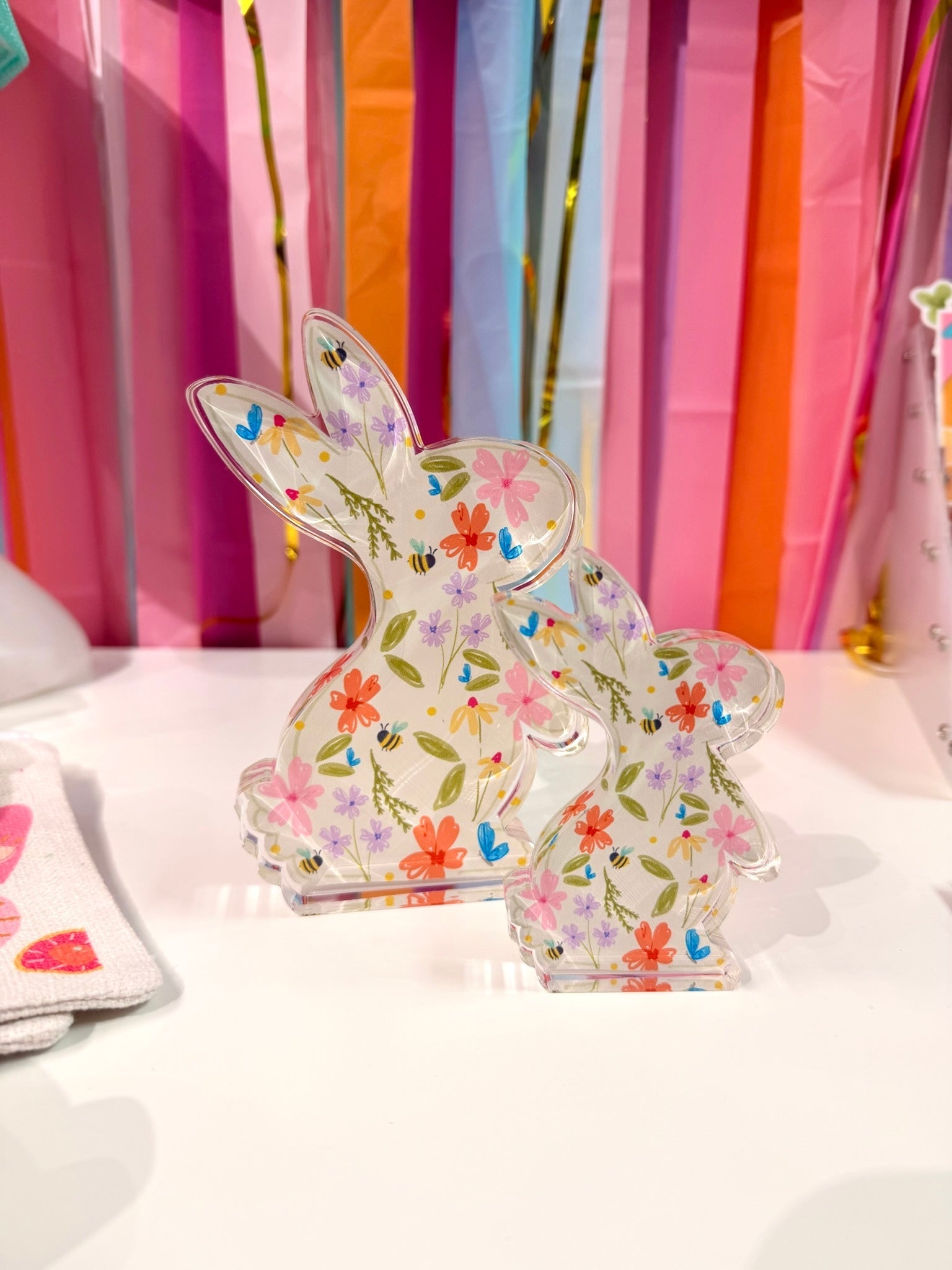 Doodles by Rebekah floral bunny acrylic home decor featuring whimsical hand-painted spring wildflowers and honeybee patterns. These colorful Easter tabletop decorations are shown against a bright party streamer background.