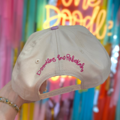 Person holding a cap with text in front of colorful decorations and a neon sign.