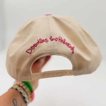 Beige cap with pink text held by a person against a white background