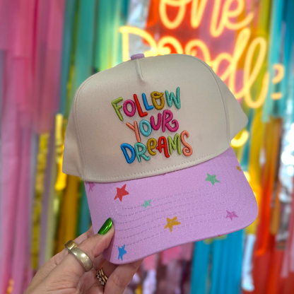 Cap with 'Follow Your Dreams' text held in front of colorful balloons and 'One Doodle' sign.