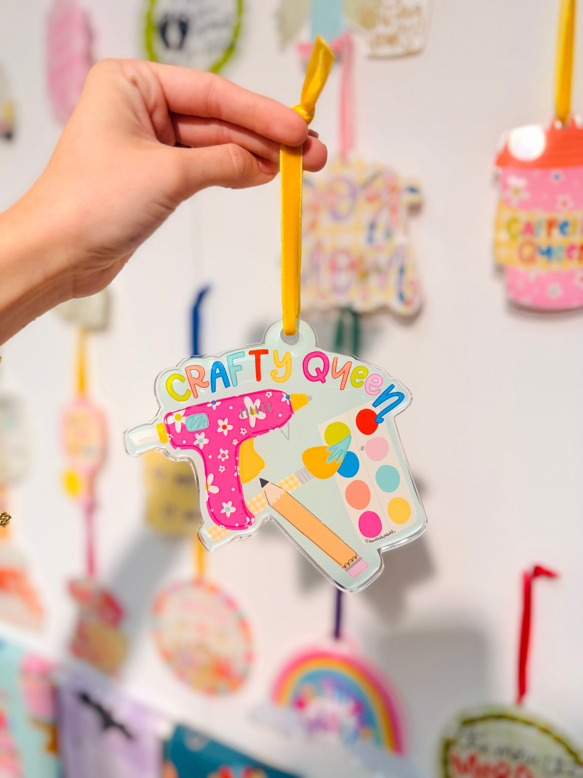 Colorful 'Crafty Queen" ornament held by a hand with a blurred background of similar decorations.