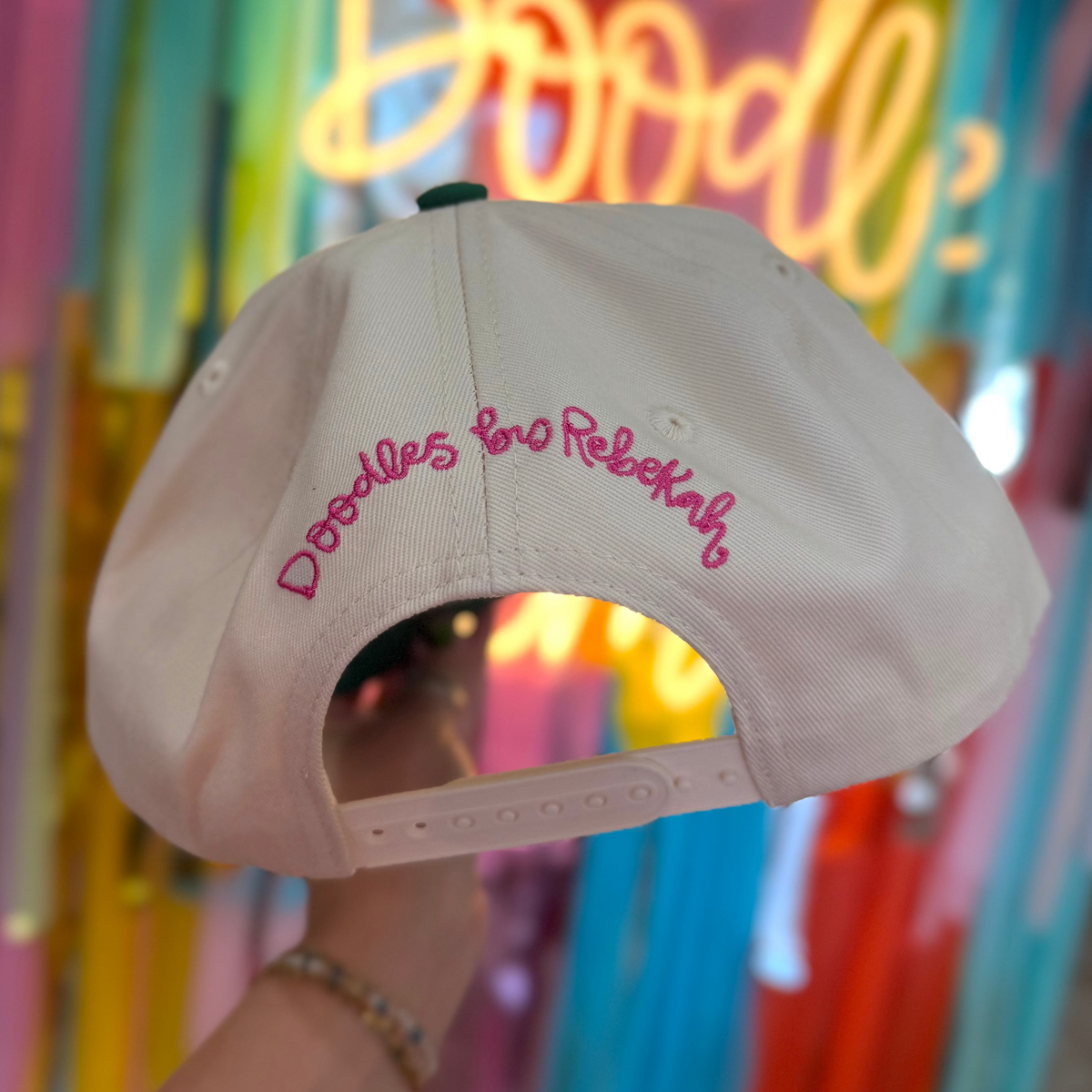 Beige cap with pink text held in front of a colorful, illuminated 'One Doodle' sign.