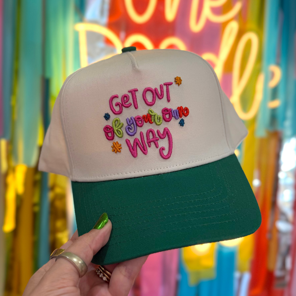 Cap with 'Get Out Be Your Own Way' text held in front of colorful streamers and a neon sign.