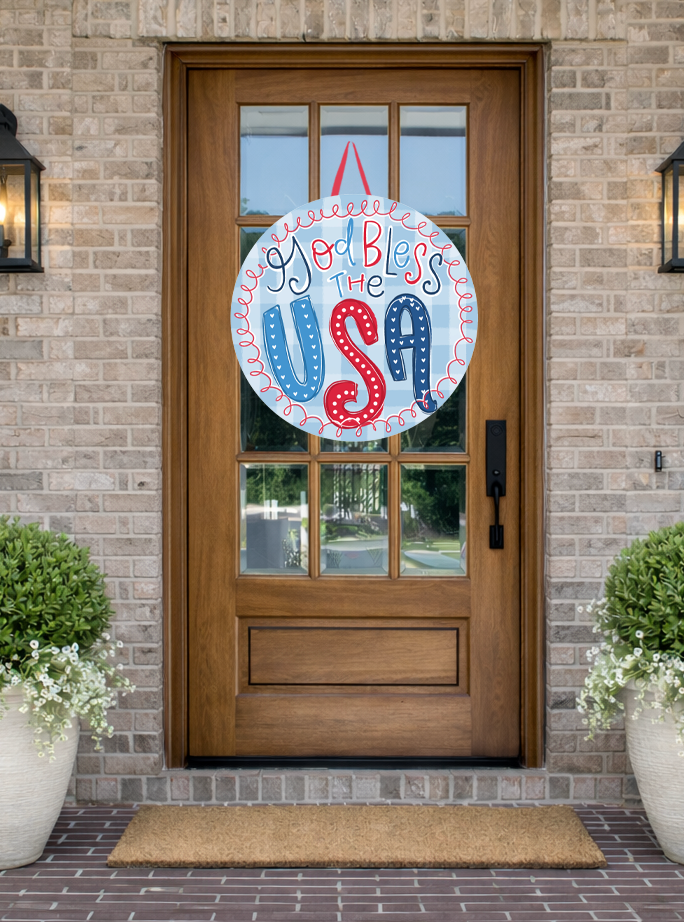 Doodles by Rebekah patriotic "God Bless the USA" door decor featuring red, white, and blue hand-lettering on a rustic front porch.