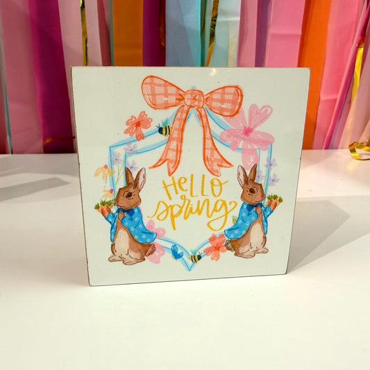 Doodles by Rebekah "Hello Spring" wooden block home decor featuring whimsical hand-painted rabbits in blue coats and garden-themed accents. This colorful seasonal tabletop or shelf decoration is shown against a bright party streamer background.