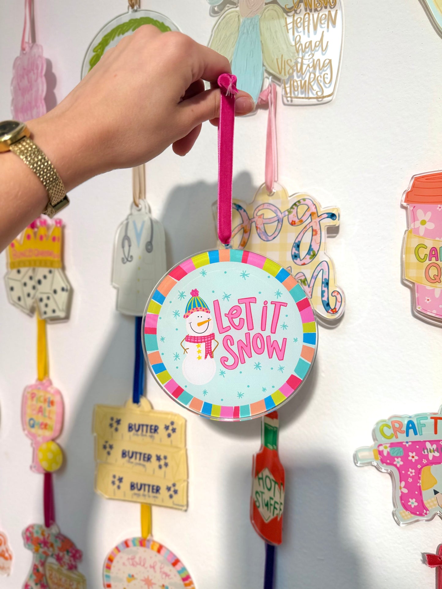 Hand holding a colorful 'Let It Snow' ornament against a white background with other decorative items.