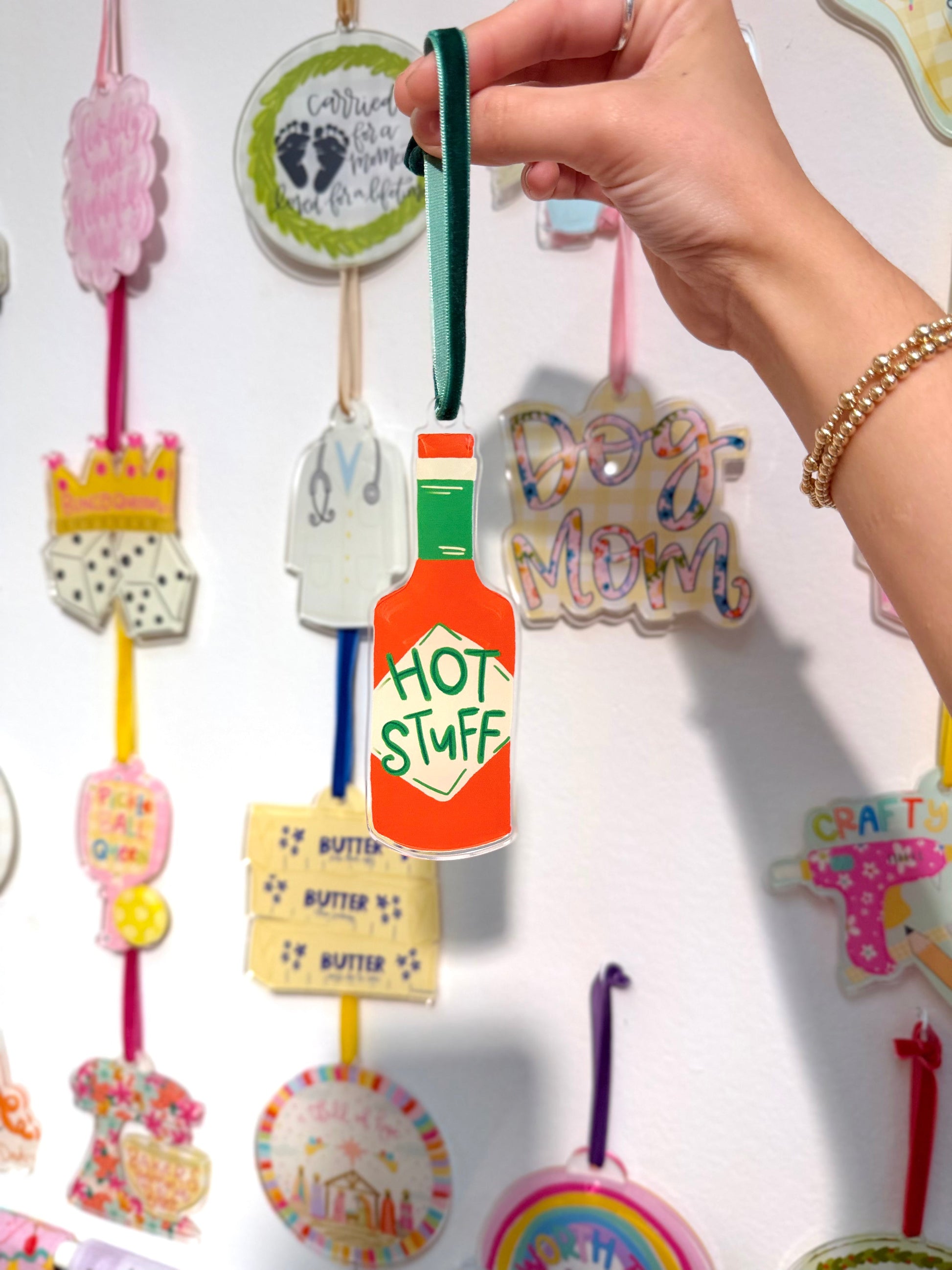 Hand holding a colorful hot sauce ornament with 'Hot Stuff' design against a wall of various ornaments