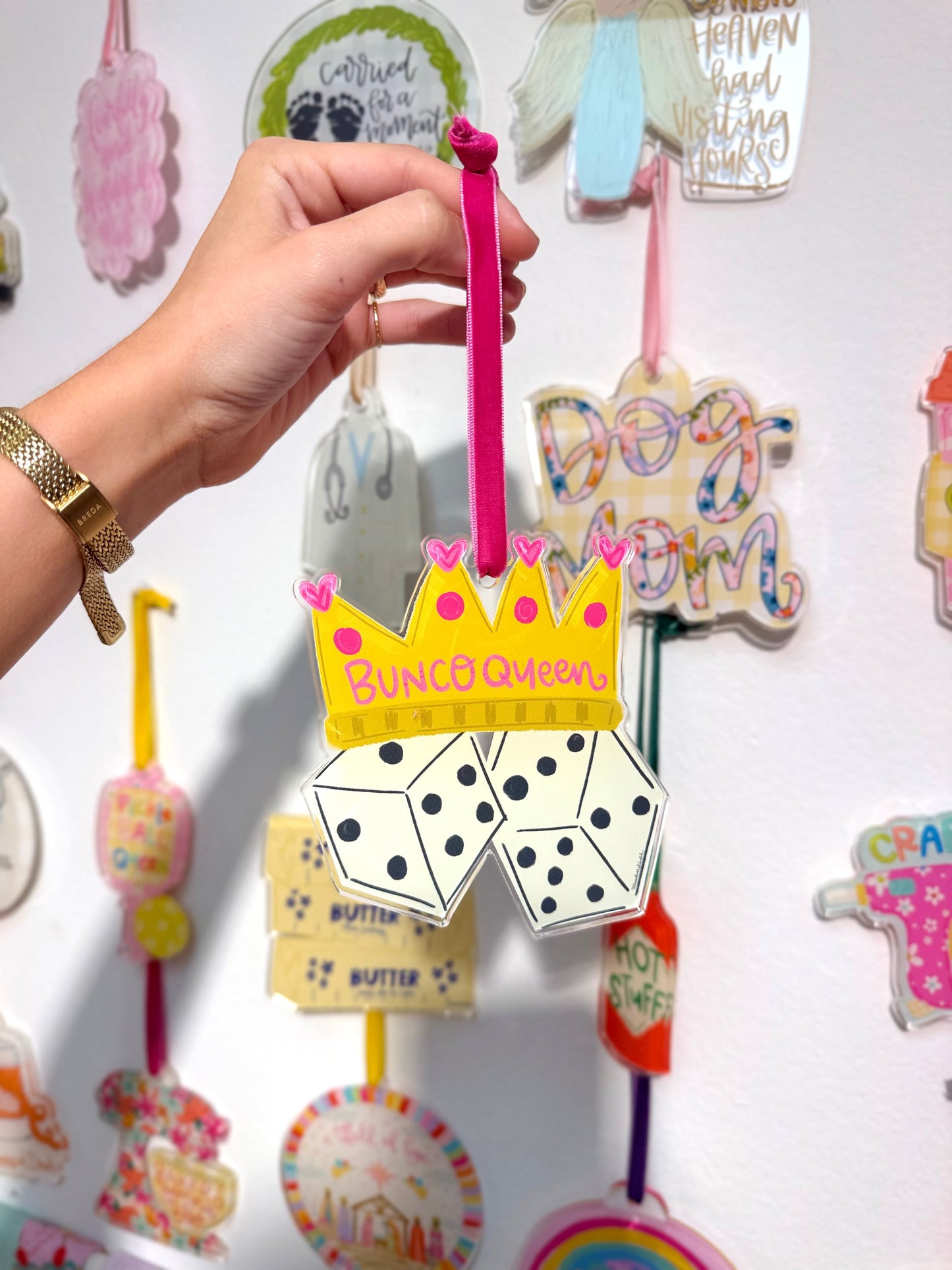 Hand holding a colorful ornament with text "Bunco Queen" and graphics against a wall of ornaments