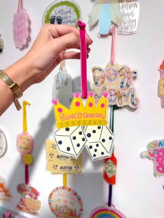 Hand holding a colorful ornament with text "Bunco Queen" and graphics against a wall of ornaments