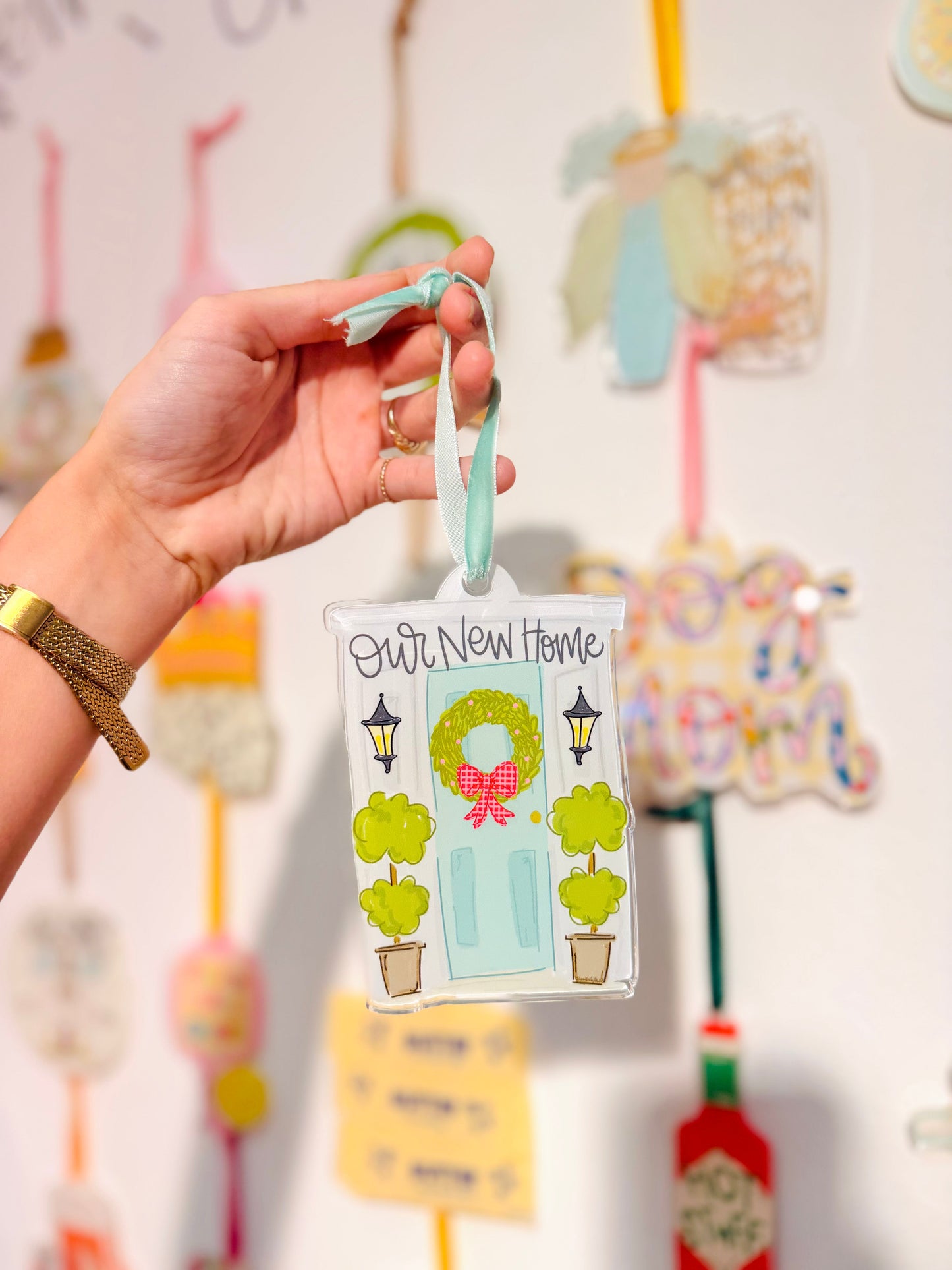 Hand holding a decorative New Home Ornament with a house and wreath design against a colorful background.