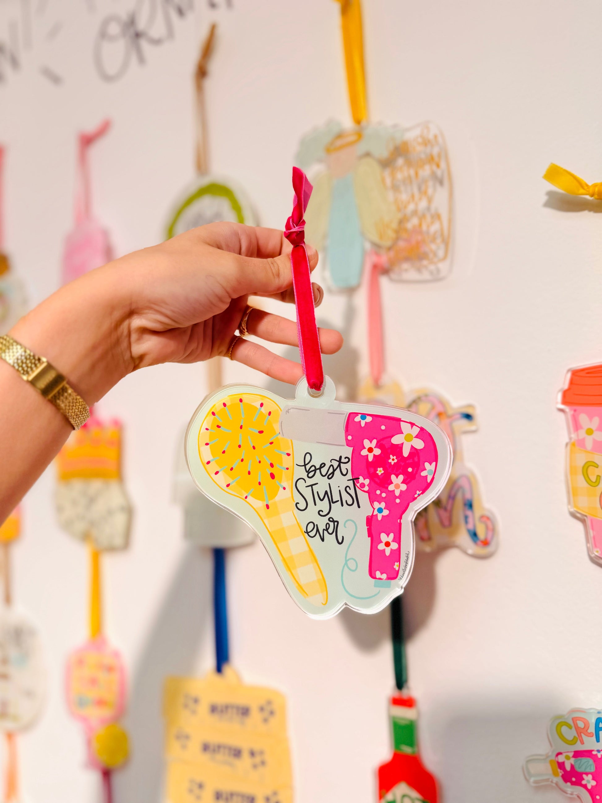 Decorative hair dryer-shaped ornament with 'Best Stylist Ever' text, held against a wall with similar ornaments.