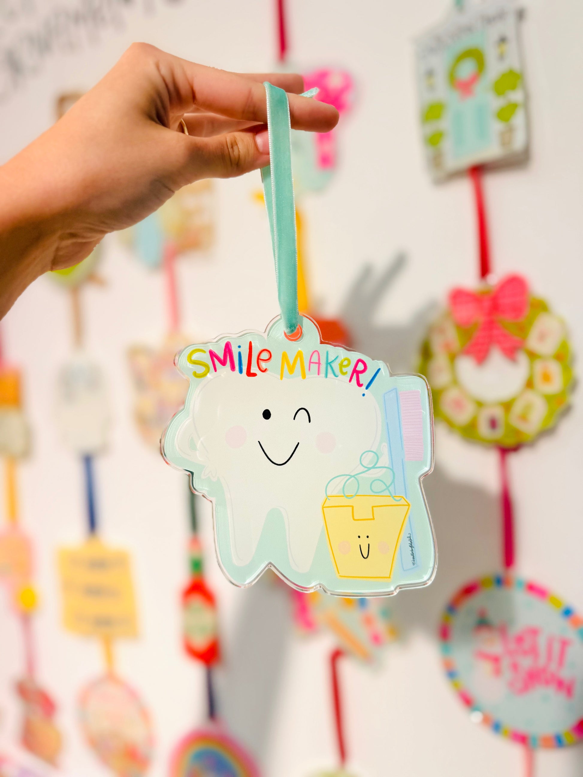 Hand holding a decorative ornament with 'Smile Maker' text and a smiley face design against a blurred background of similar decorations.