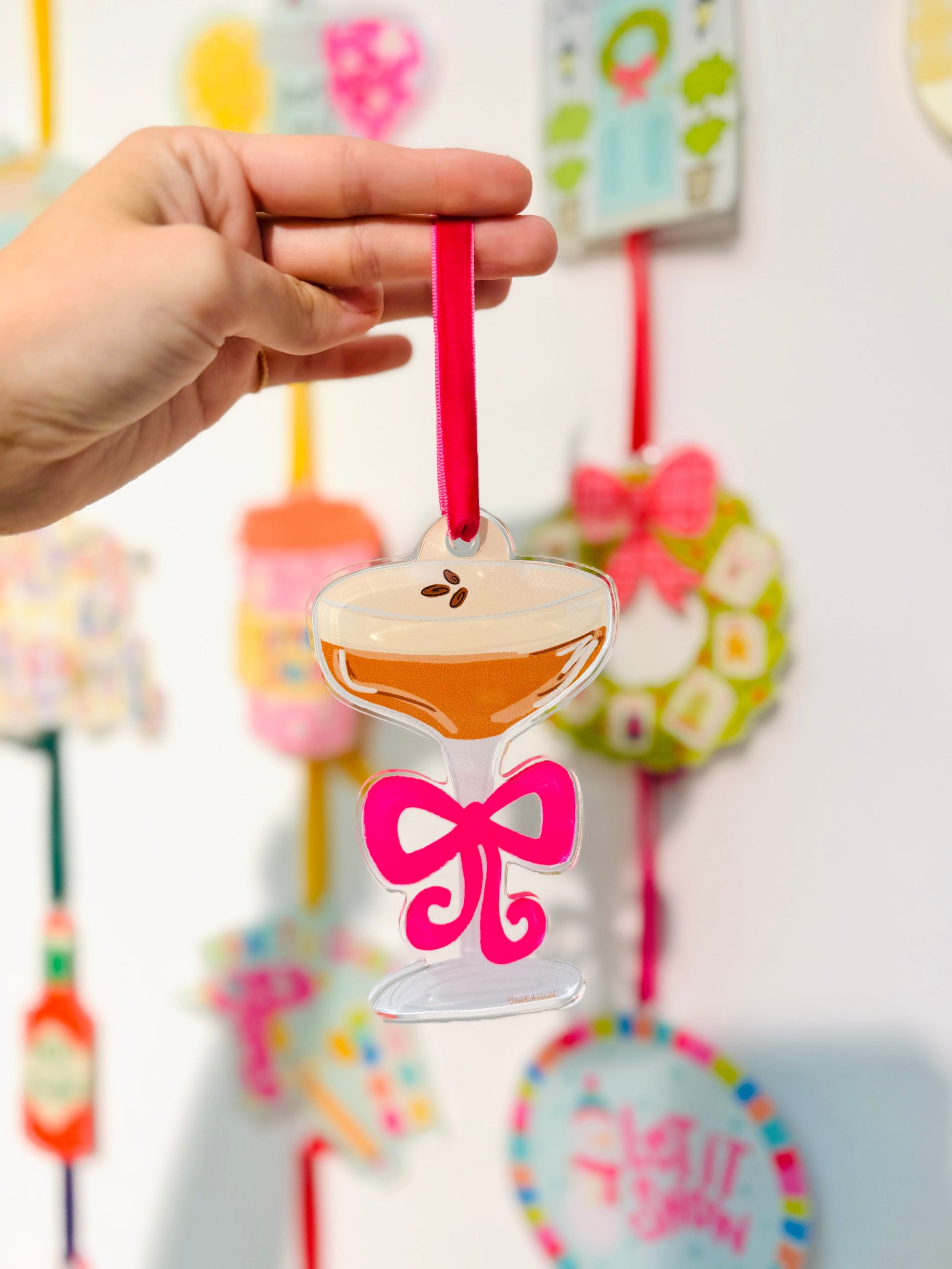 Decorative ornament shaped like a glass with a pink bow, held by a hand against a colorful background.