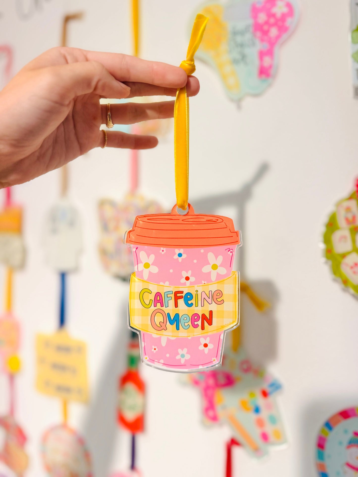 Pink drink cup ornament with 'Caffeine Queen' text held by a hand against a colorful background.
