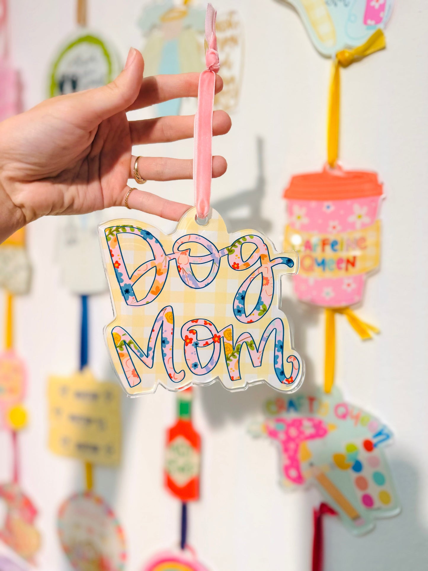 Colorful 'Dog Mom' ornament held by a hand with a blurred background of similar decorative items.