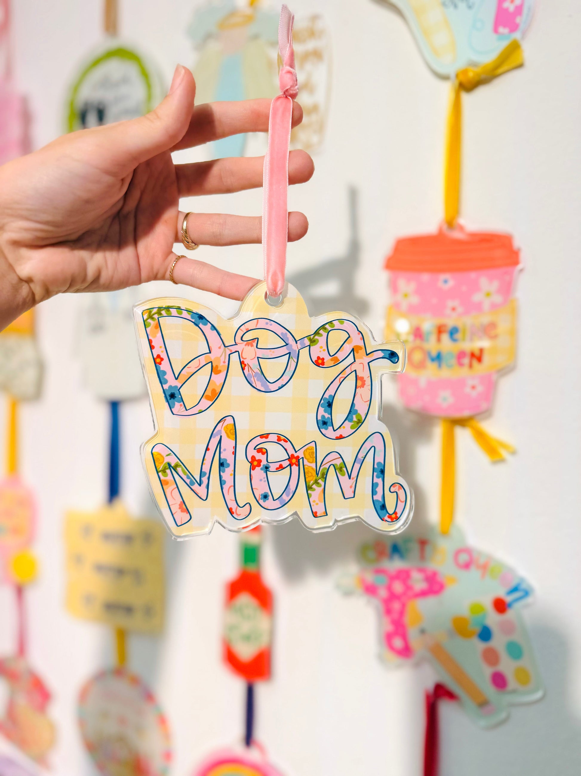 Colorful 'Dog Mom' ornament held by a hand with a blurred background of similar decorative items.
