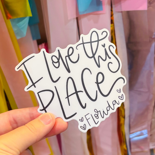 a hand holding a black and white sticker that reads "I Love This Place Florida"