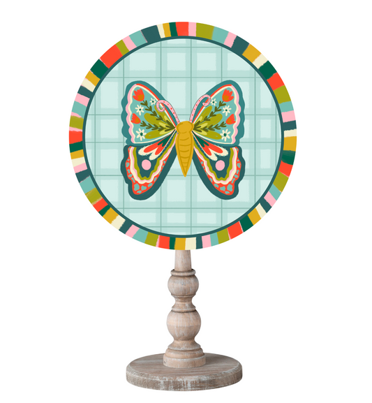 A circle topper with a colorful butterfly with floral details in it's wings with a colorful border on top of a wooden dowel