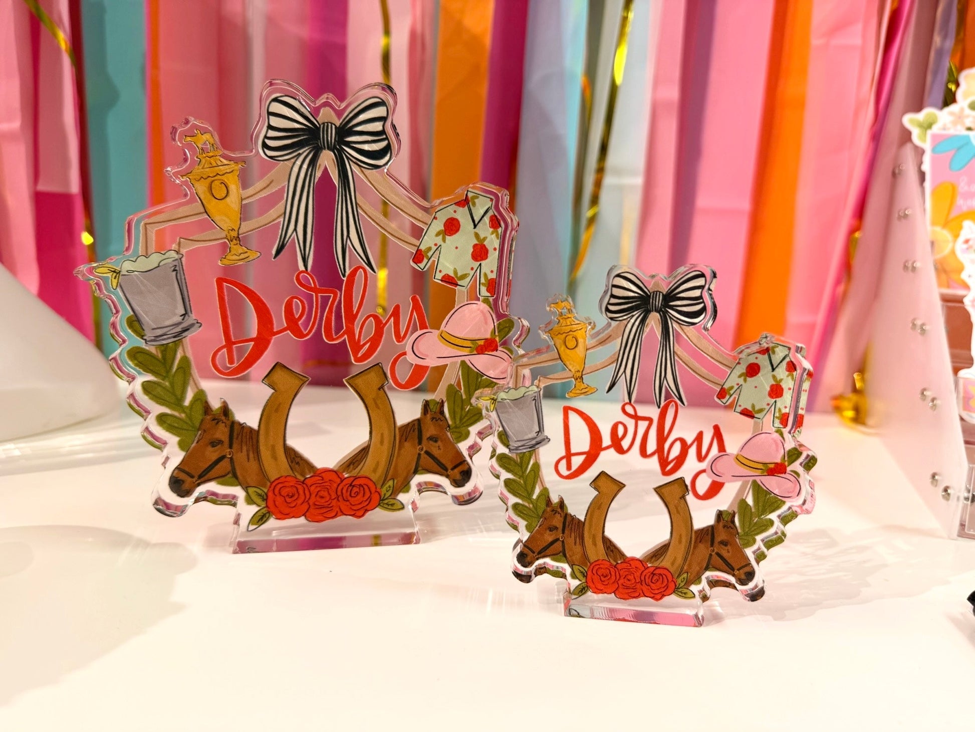 Doodles by Rebekah Kentucky Derby acrylic home decor featuring whimsical hand-painted horse racing icons, red roses, and "Derby" typography. These colorful horse racing accents are shown against a bright party streamer background.