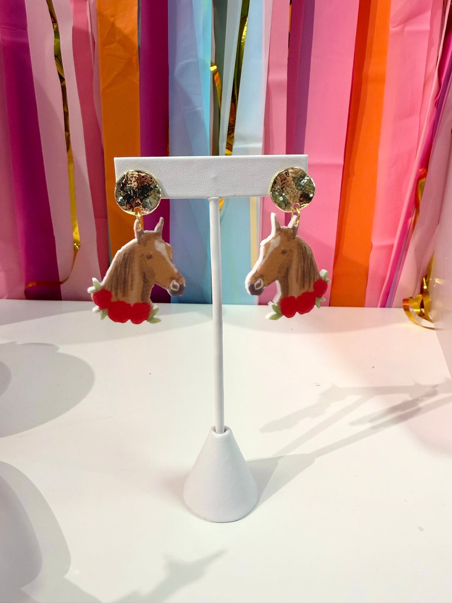 Doodles by Rebekah Kentucky Derby horse earrings featuring hand-painted thoroughbred designs, red roses, and hammered gold studs. These whimsical equestrian-themed accessories are shown against a bright party streamer backdrop.