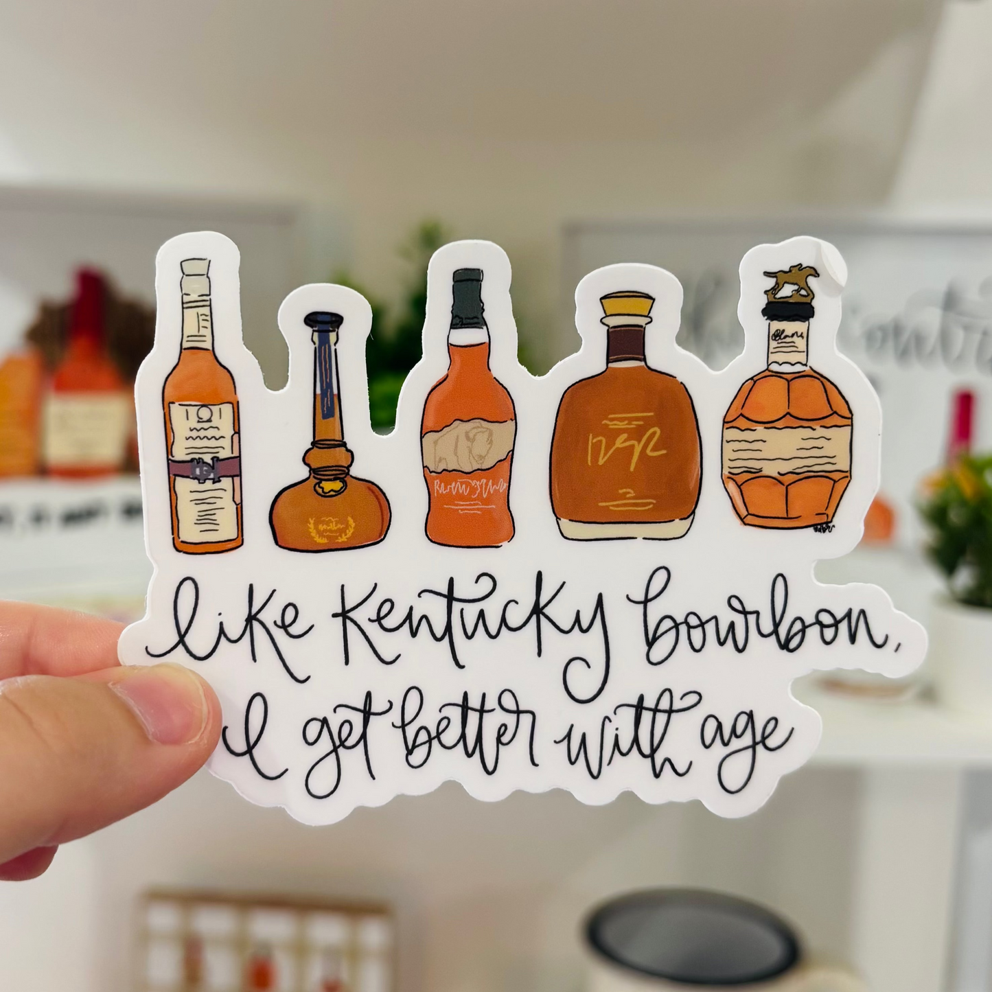 a hand holding a sticker that reads "Like Kentucky Bourbon I get better with age" with hand drawn bourbon bottles