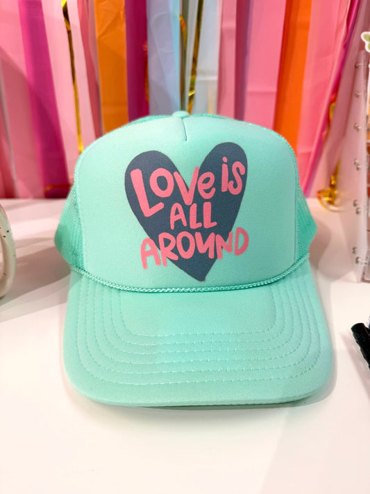 Mint green "Love Is All Around" trucker hat with a navy heart and pink lettering, featuring a whimsical hand-drawn style for Valentine's Day or everyday wear.