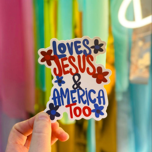 Person holding a red, white and blue sticker with flowers and the phrase "Loves Jesus & America Too"