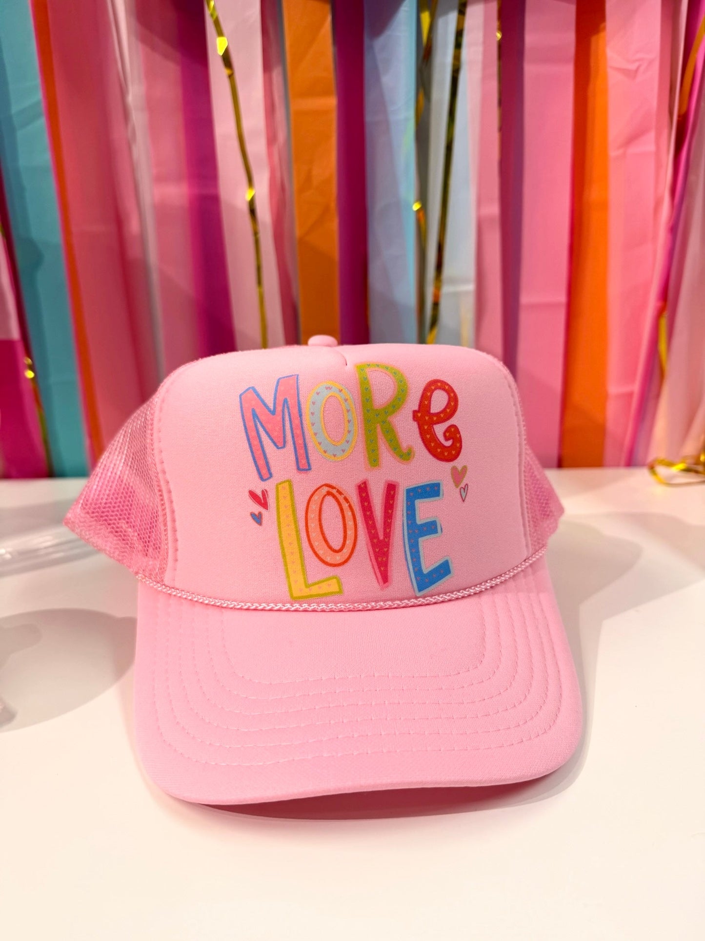 Pink "More Love" trucker hat from Doodles by Rebekah, featuring whimsical hand-drawn typography and heart illustrations on a colorful party streamer background.